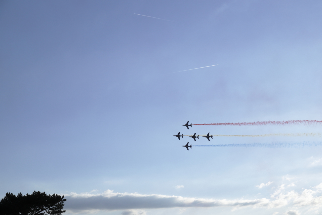 Cinq appareils en formation de la patrouille de France avec des fumigènes bleus, blancs et rouges.