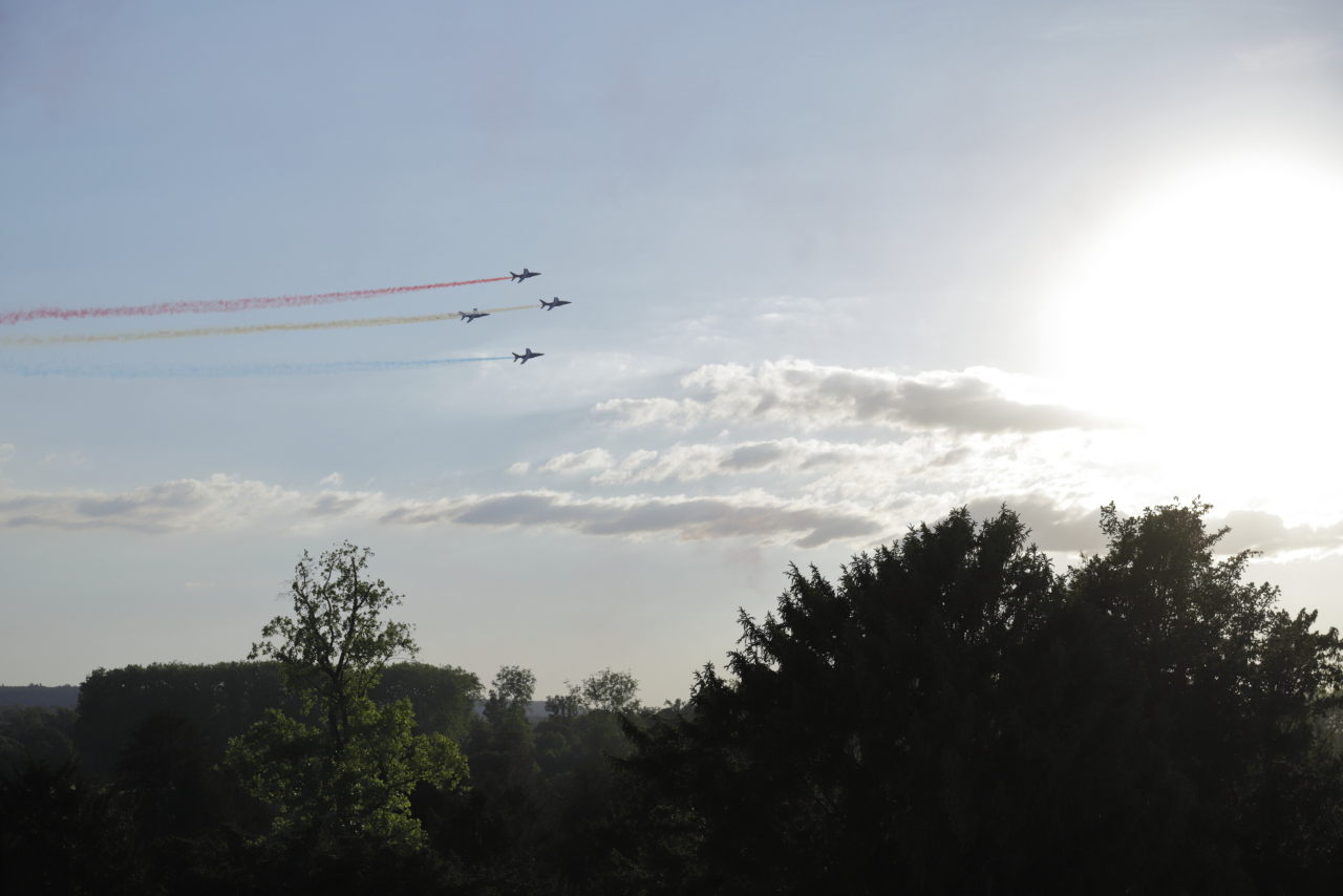 Cinq appareils en formation de la patrouille de France avec des fumigènes bleus, blancs et rouges.