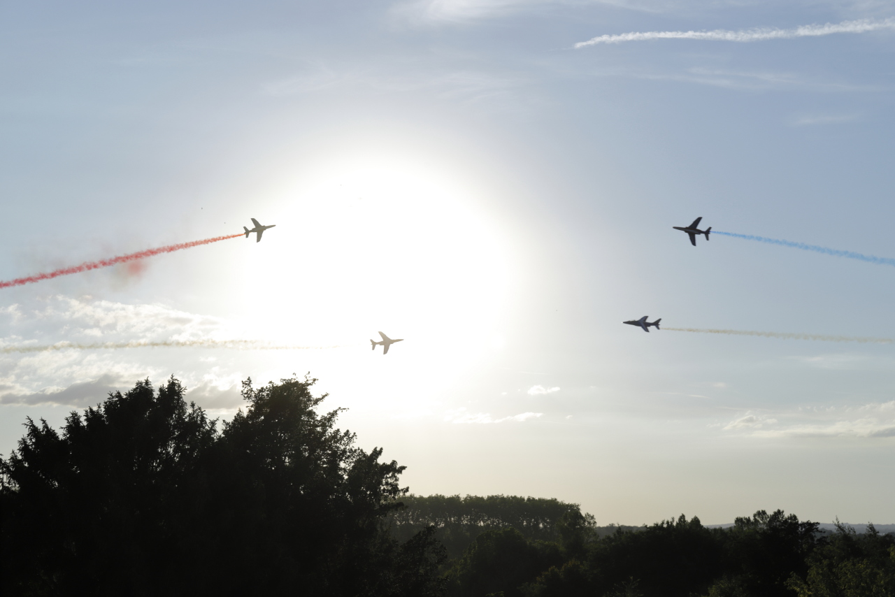 Quatre appareils de la patrouille de France qui se croisent face au soleil avec des fumigènes bleus, blancs et rouges.