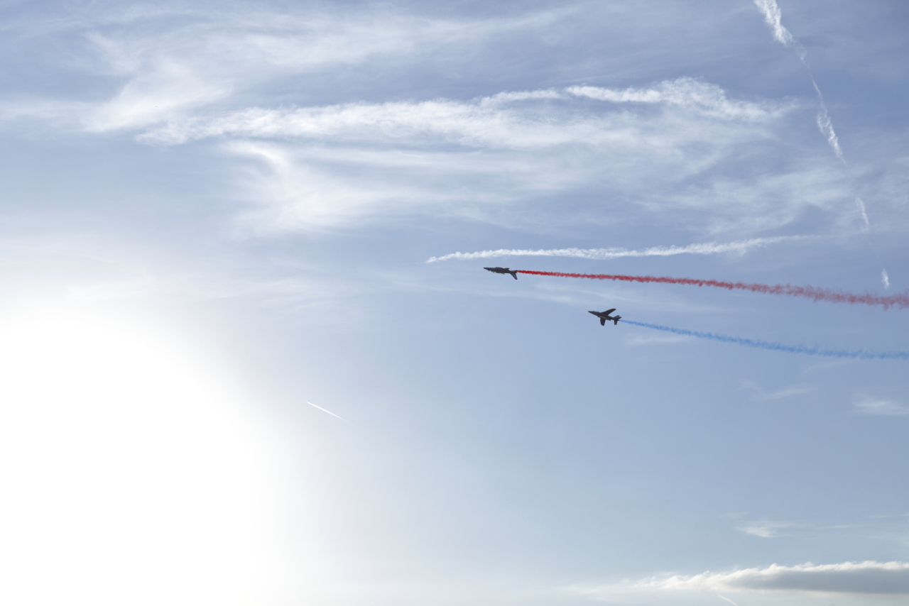 Deux appareils de la patrouille de France qui se suivent, l’un avec un fumigène rouge et l’autre avec un bleu.