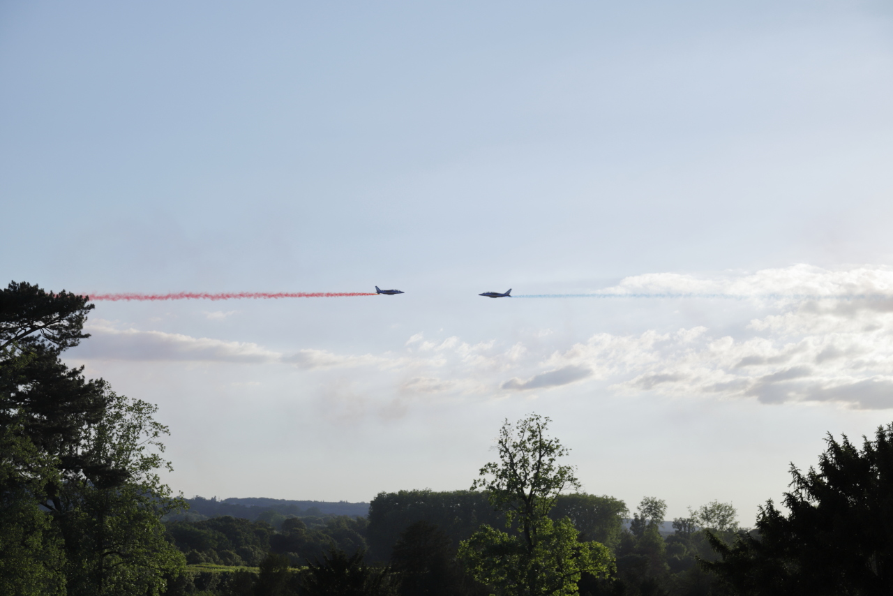 Deux appareils de la patrouille de France se faisant face devant le soleil, l’un avec un fumigène rouge et l’autre avec un bleu.